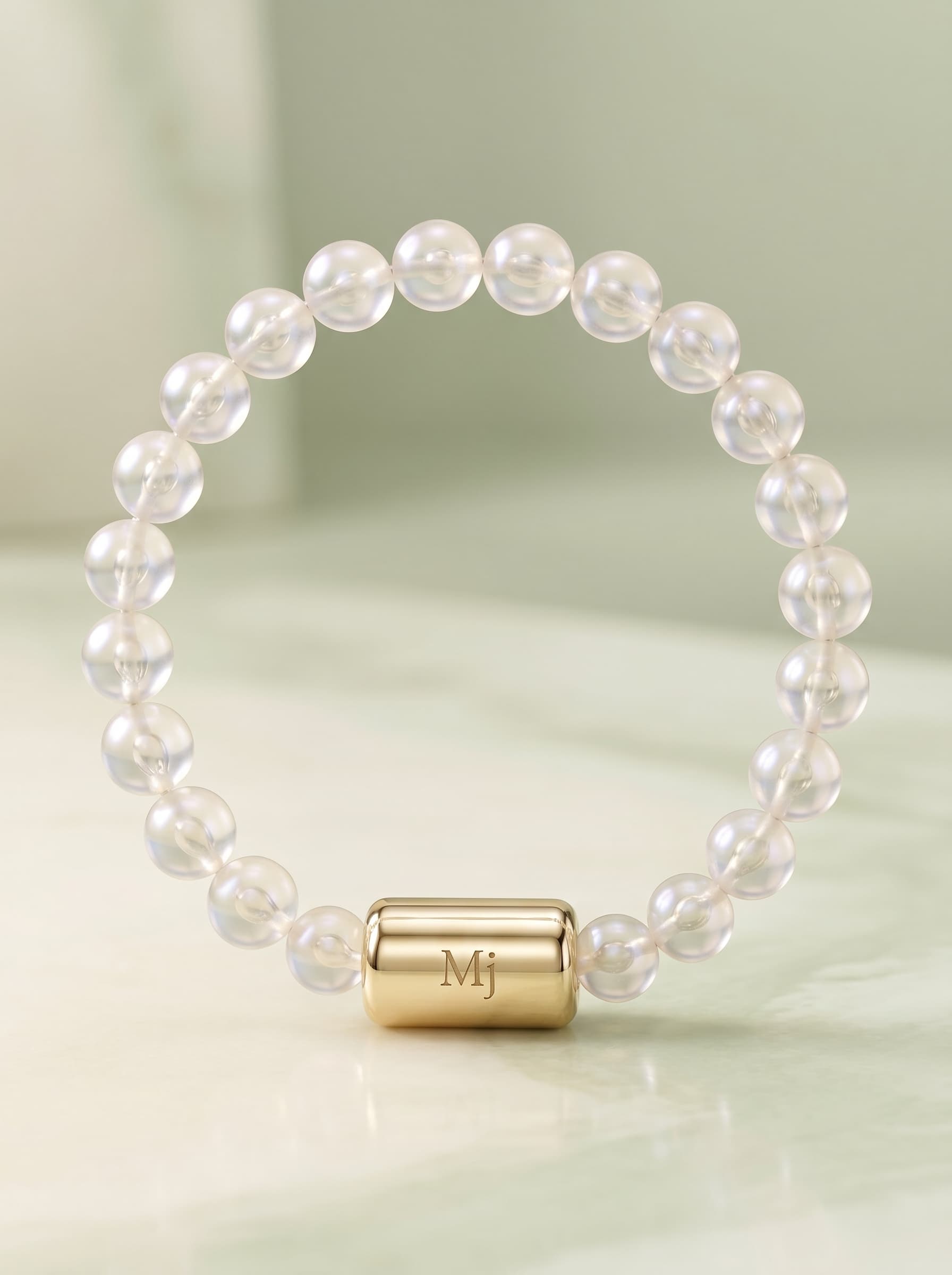 The MEDIJEWEL bracelet laid in a circle: clear frosted beads with an engraved cylindrical clasp marked Mj, on a soft sage surface.